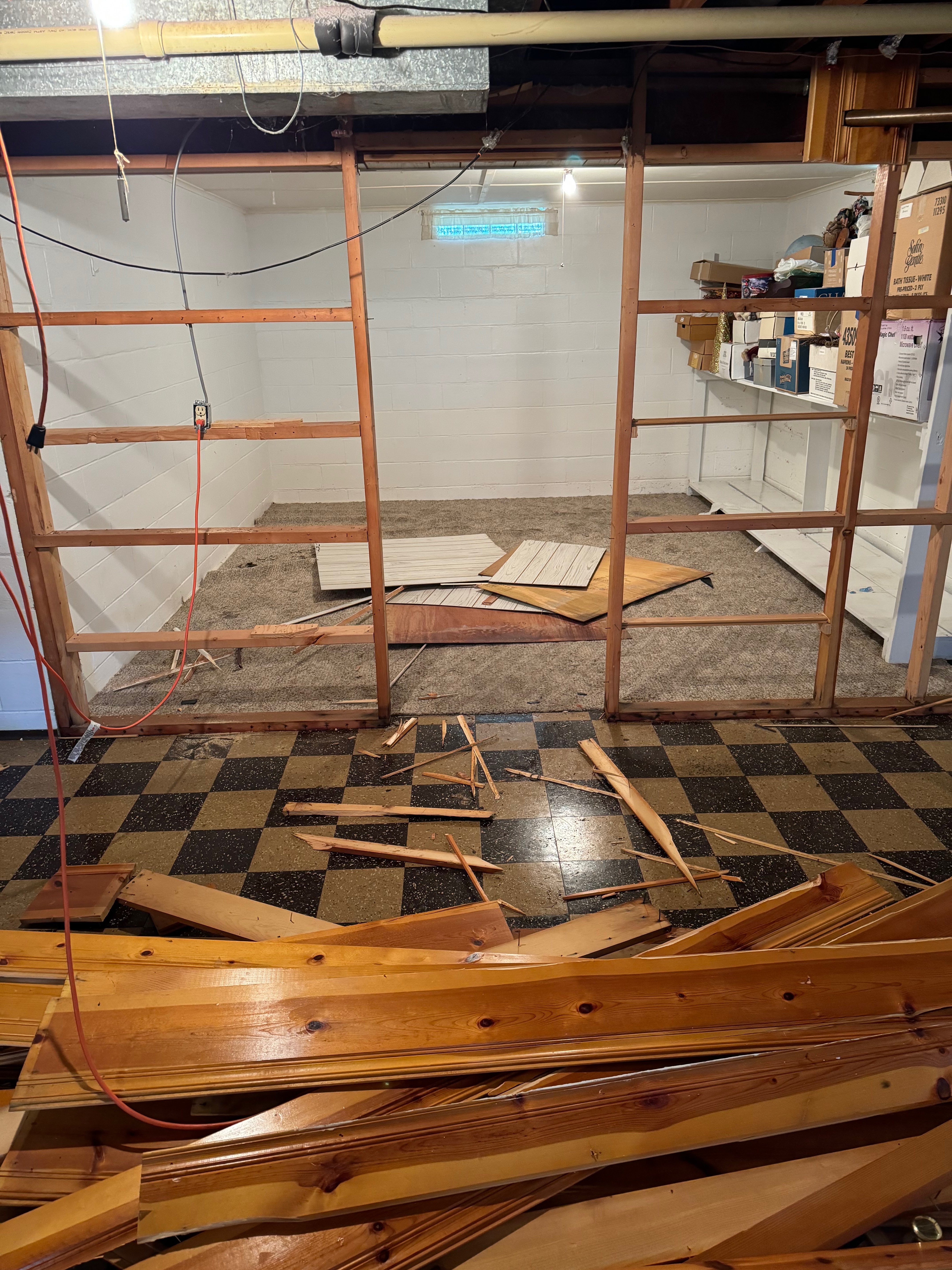 Basement area with exposed wooden framing and scattered damaged wood pieces on checkered floor indicating structural water damage requiring remediation