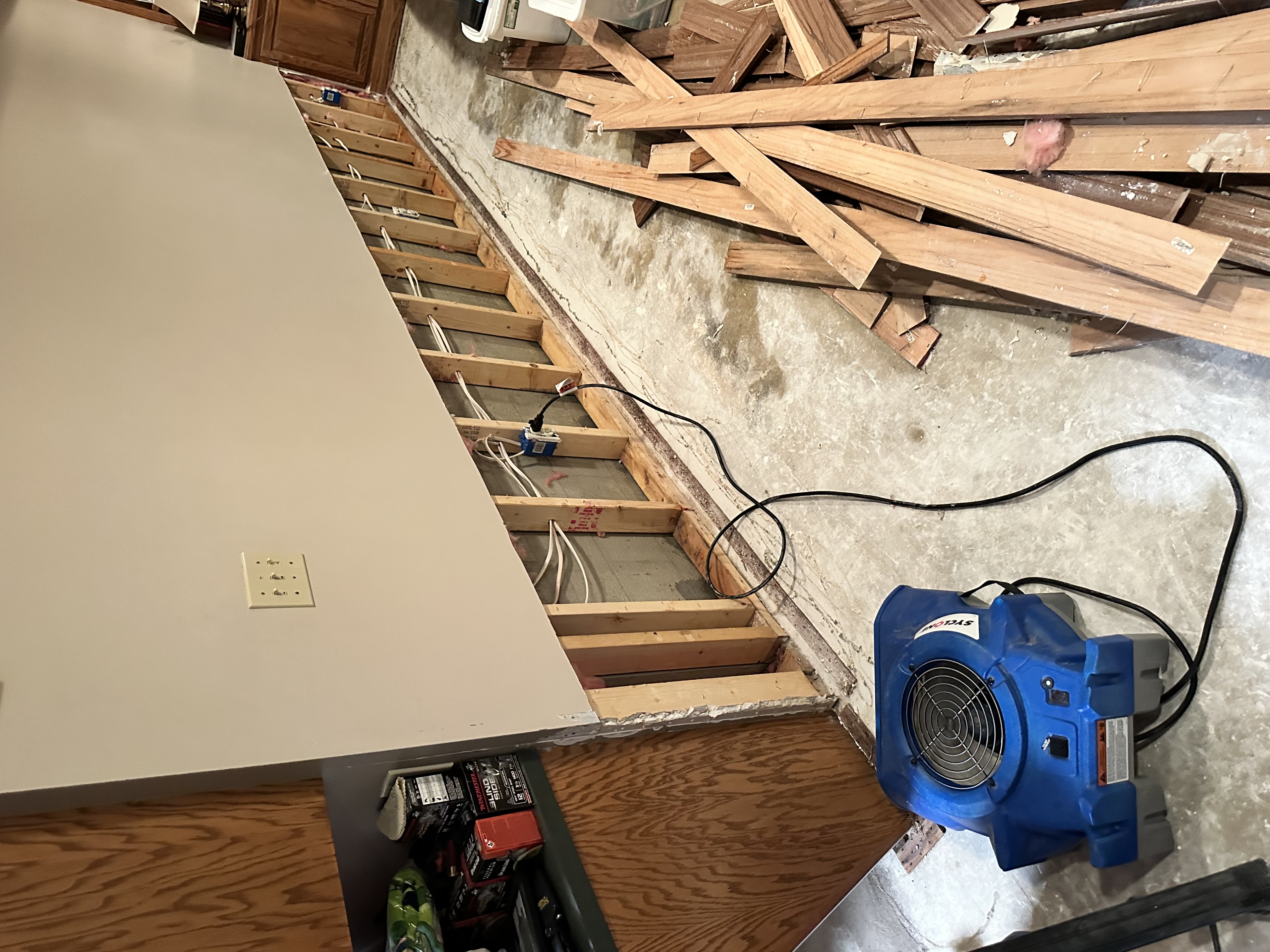 Basement water damage restoration with exposed wall framing, blue air mover fan on floor, and scattered wooden materials showing water damage to concrete surfaces